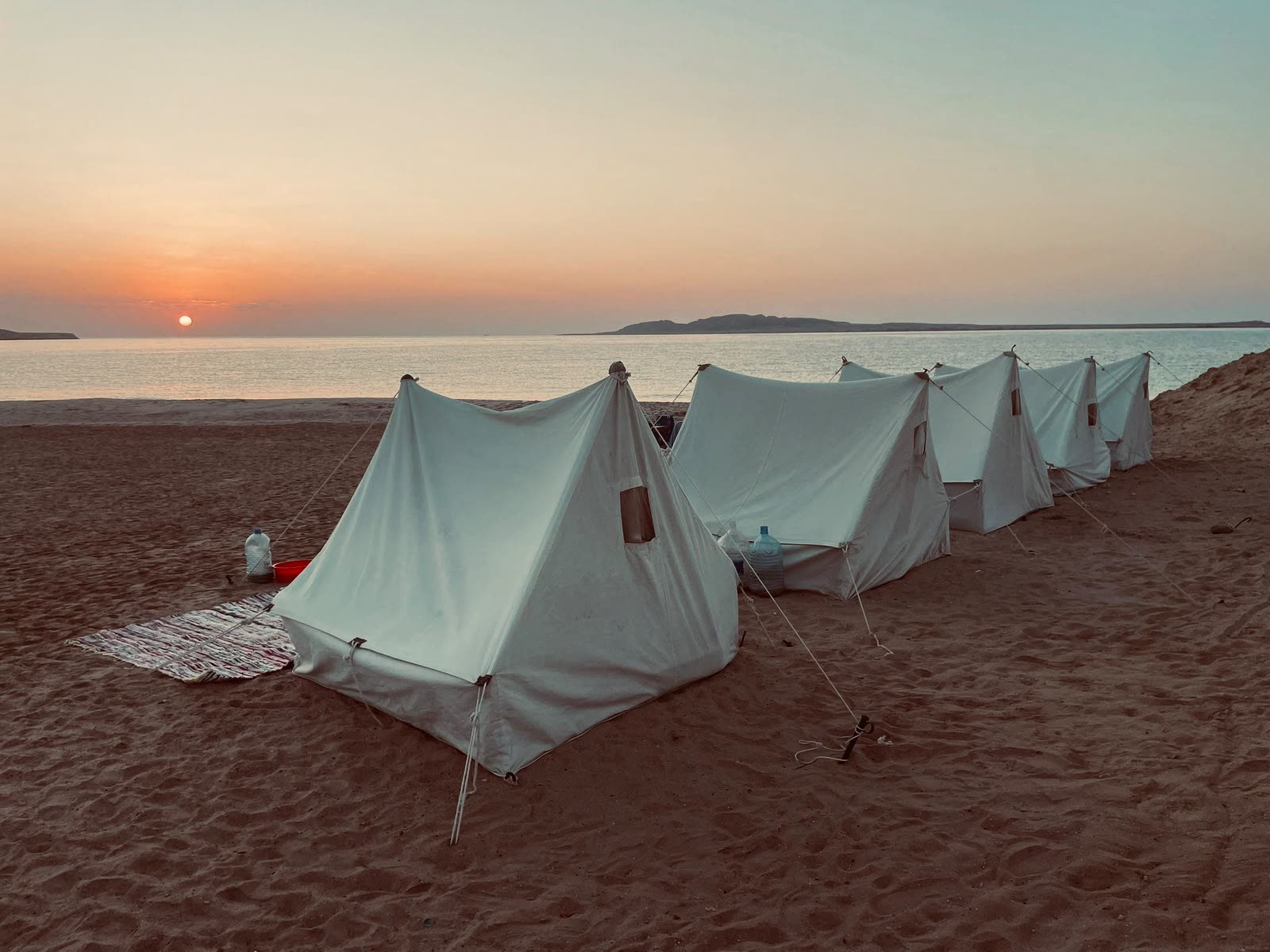 A row of white canvas tents on the sand at sunset, calm sea and a low island behind.