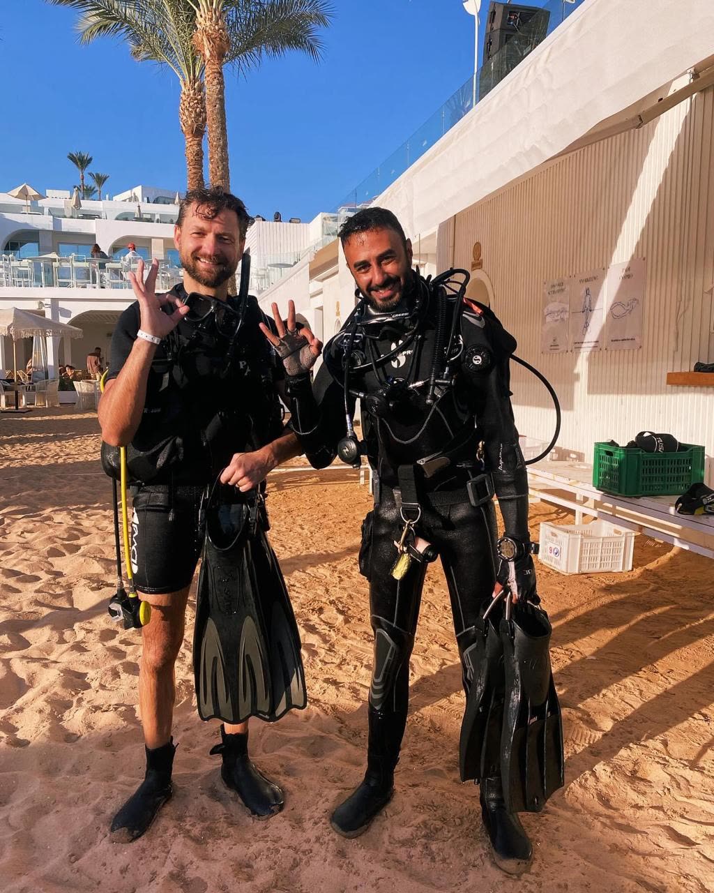 Two divers standing on the sand in wetsuits and BCDs, fins in hand.