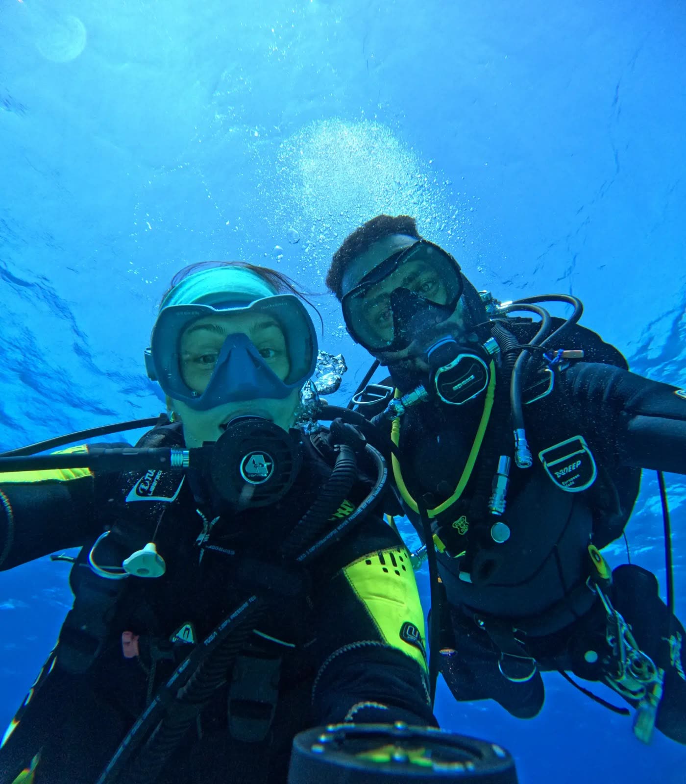 Two divers taking a selfie underwater, sunlight and bubbles above.