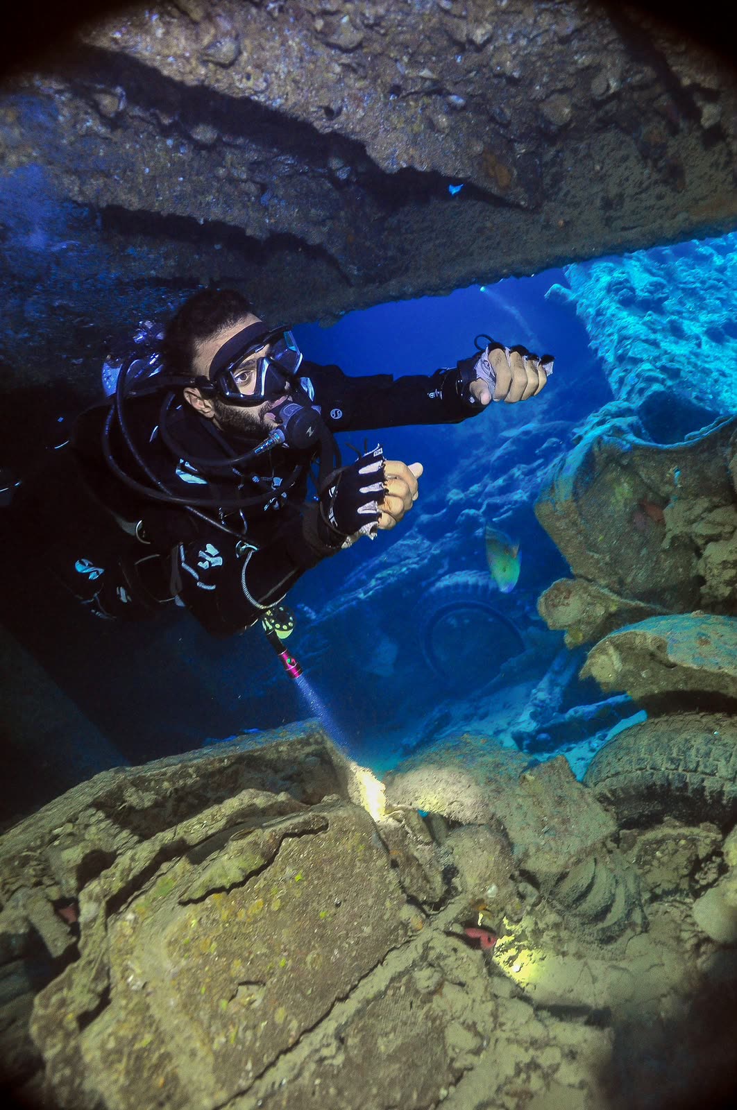 A diver with a torch inside the wreck, cargo in the hold below.