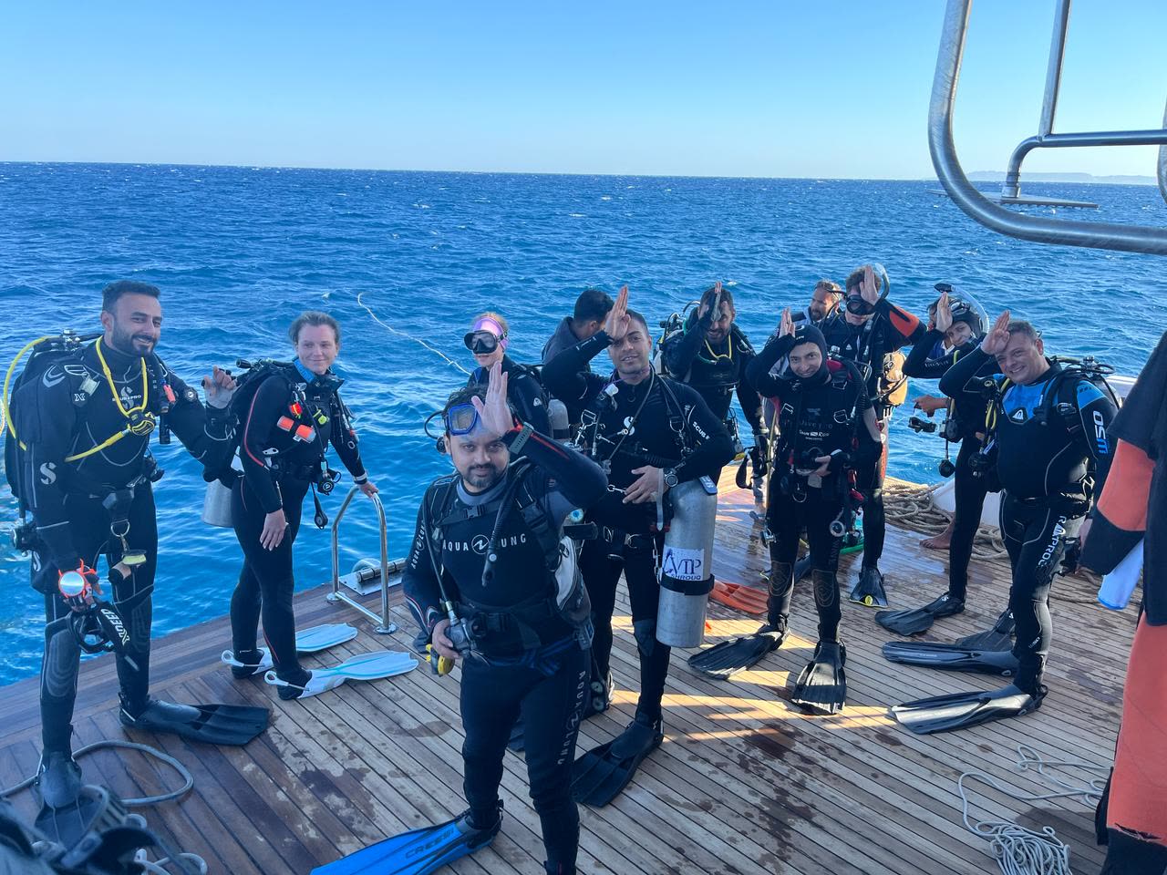 A dozen divers kitted up on deck, fins at their feet, flat blue water behind them.